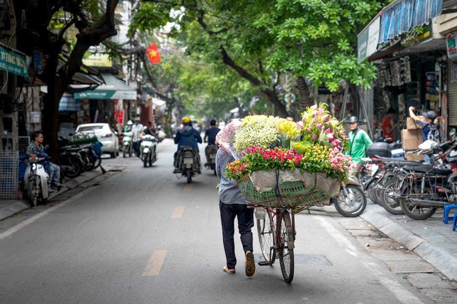 A street scene featuring a flower seller pushing a bicycle loaded with a large bouquet of fresh flowers, including pink roses, white hydrangeas, and green foliage, wrapped in decorative paper and ribb