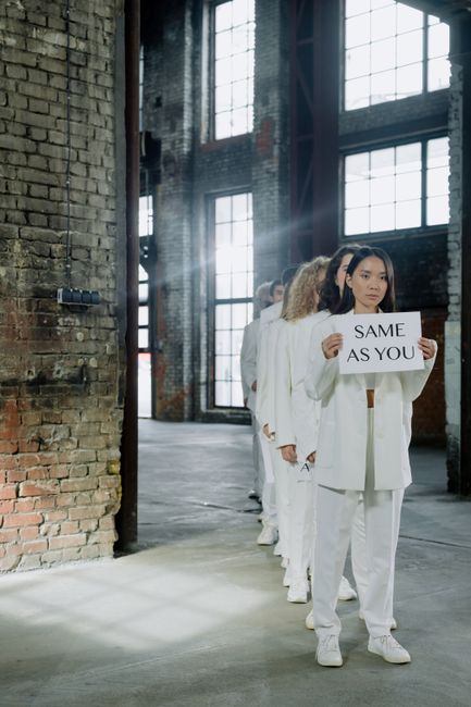 A line of women dressed in white suits standing inside an industrial-style space with exposed brick walls and large windows, holding a sign that reads 'SAME AS YOU'. The natural light streaming throug