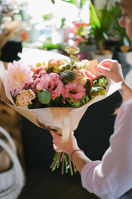 A person holding a large, hand-tied bouquet of fresh flowers in a floral wrap of cream and white paper. The arrangement includes soft pink peonies, white carnations, and small purple and white filler 