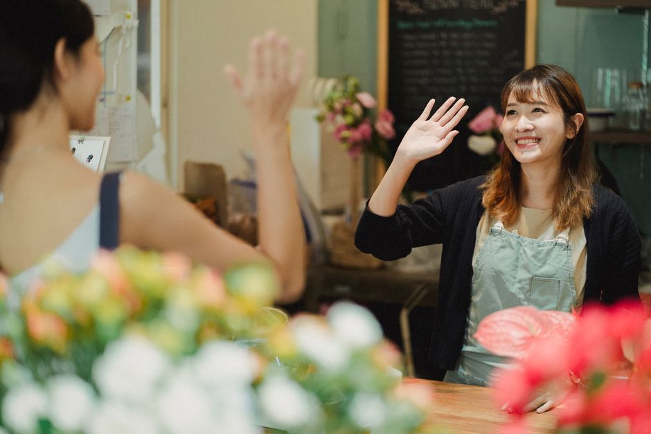 A smiling young woman with shoulder-length brown hair, wearing a light-colored apron over a beige top and a black cardigan, is engaged in a friendly high-five with a customer at a flower shop. The sho