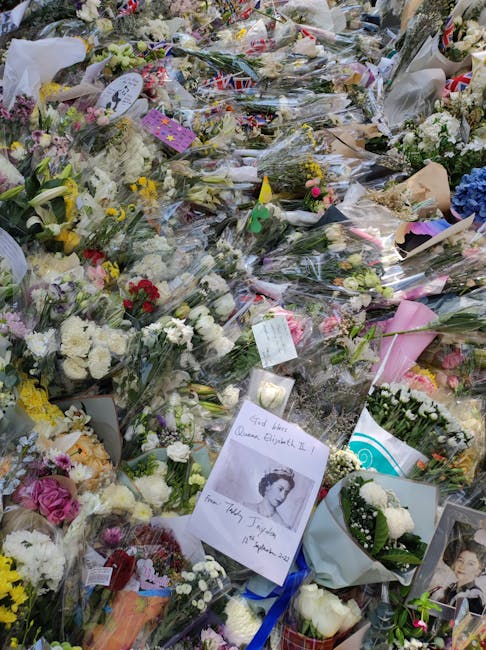 A densely packed floral tribute scene featuring a wide variety of fresh flowers arranged in bouquets and wreaths. The arrangement includes roses, carnations, lilies, and chrysanthemums in soft pastel 