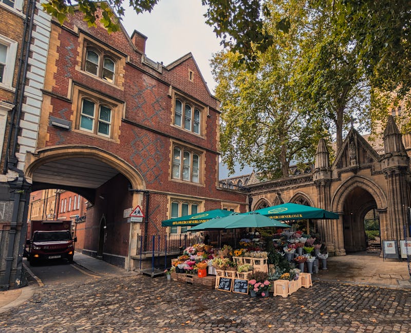 A flower stall situated on a cobblestone street near the historic, red-brick architecture of Sloane Square in Chelsea. The stall, protected by a green umbrella bearing the floristchelsea.org.uk brandi