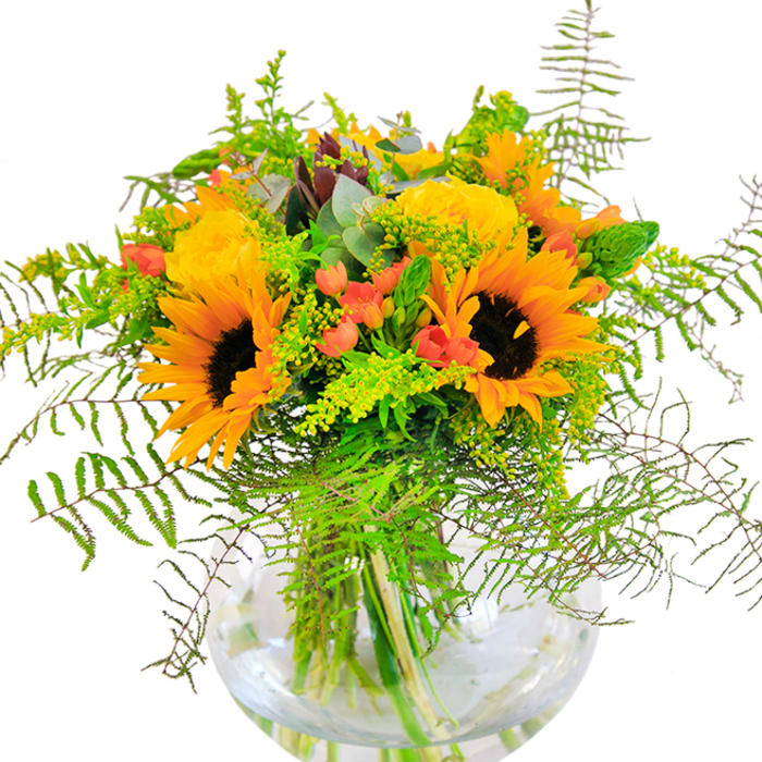 Evoking the sophisticated yet relaxed spirit of Chelsea, this opulent bouquet is a sunlit embrace in floral form. Dominating the scene are bold, golden sunflowers, their faces aglow and edged with bright yellow-orange petals, conjuring images of riverside strolls near Chelsea Bridge. These sunny blooms are complemented by graceful yellow garden roses, each petal intricately folded in a classic spiral, recalling the refined gardens that dot this iconic London area. Interwoven throughout, gleaming clusters of deep red Hypericum berries sparkle against a verdant backdrop, providing a lively contrast and a dash of bold colour, like the vibrant finds at a Chelsea Flower Market stall. Textural surprises abound, with soft, feathery ferns and wispy green fillers, such as solidago, tumbling gently, recalling tranquil moments in Chelsea Physic Garden. Delicate bluish-green eucalyptus leaves emerge subtly between blooms, lending a rustic, contemporary edge. Every stem is anchored securely in a round, fishbowl-style glass vase, the glistening water magnifying the stems' intricate network and reflecting soft daylight. The bouquet radiates warmth-a gentle rose aroma mingles with fresh greenery, carrying the senses to a sun-warmed terrace. This arrangement is ideal for celebratory gatherings, birthdays or simply to add a burst of optimistic beauty to a Chelsea living room, expertly arranged by a local florist who knows just how to express the neighbourhood's unique charm through flowers.