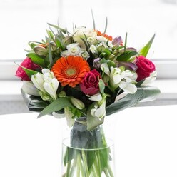 Echoing the timeless elegance of Chelsea, this visually stunning bouquet is a testament to floral artistry and vibrant local character. Dominating the composition are four gerbera daisies-two in brilliant orange, blazing with the zest of autumn leaves along Burton Court, and two in creamy peach with warm beige undertones, offering a gentle, embracing glow. Deep magenta roses are artfully nestled among the daisies, their lush spirals hinting at the romantic courtyards tucked behind Chelsea's stately homes. A scattering of white alstroemeria and delicate white spray roses catches the natural light, bringing luminosity and a sense of gentle movement, while subtle pink blooms and clusters of tiny white filler flowers add to the allure and generous feel of the bouquet. Glossy foliage, richly emerald, cradles the flowers, while paddle-shaped aspidistra leaves sweep upward and around the base, forming a sculpted green collar that imparts a serene yet sturdy frame reminiscent of Chelsea Embankment's manicured gardens. The bouquet's arrangement feels carefully choreographed yet effortlessly lush, with slender stems confidently displayed through the clear glass vase-a testament to both freshness and meticulous design. Soft light pools over the bouquet, evoking an air of renewal, optimism, and understated luxury. Whether used to celebrate a new home on Tite Street, honour a friend's milestone, or grace a Chelsea dining table, this bouquet encapsulates the warmth, artistry, and individuality of a local florist deeply rooted in Chelsea's vibrant community.